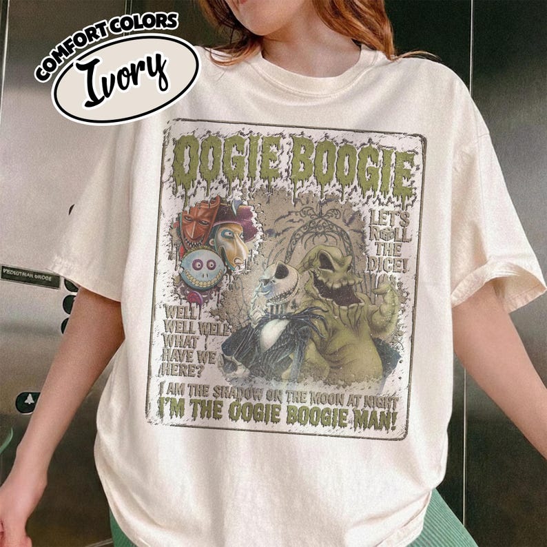 May include: Ivory-colored t-shirt featuring a graphic from "The Nightmare Before Christmas." The design includes the text "DOGIE BOOGIE" and images of characters like Oogie Boogie and Jack Skellington. The shirt has a crew neck and short sleeves.