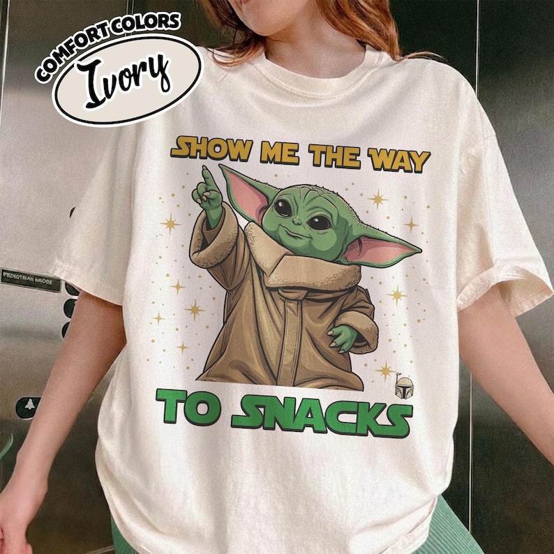 May include: Ivory-colored t-shirt with a graphic of Grogu from The Mandalorian. The image shows Grogu pointing upwards with the text "SHOW ME THE WAY TO SNACKS". The shirt has a Comfort Colors label.