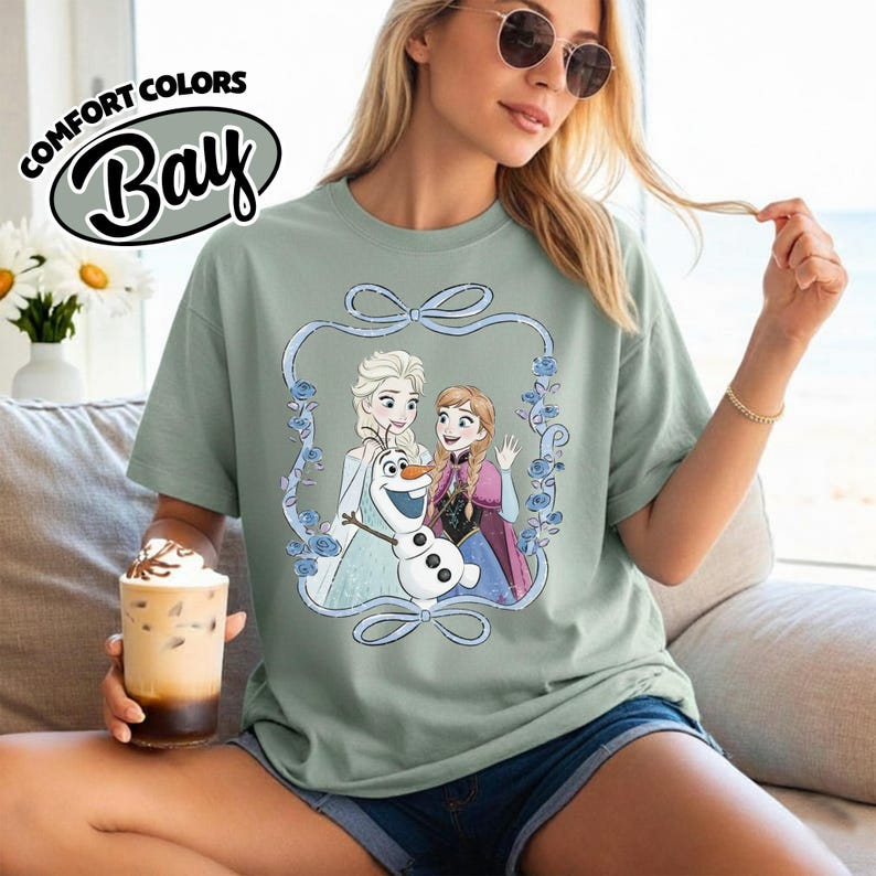 May include: A sage green t-shirt featuring a graphic of Elsa, Anna, and Olaf from Frozen, framed by a blue ribbon and floral design. The shirt has the text "COMFORT COLORS Bay" in a circular design.