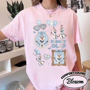 May include: Pink Comfort Colors t-shirt featuring a Disney Olaf graphic. The design includes Olaf characters, blue balloons, hearts, and the text "Disney Olaf". The shirt has a round neckline and short sleeves.
