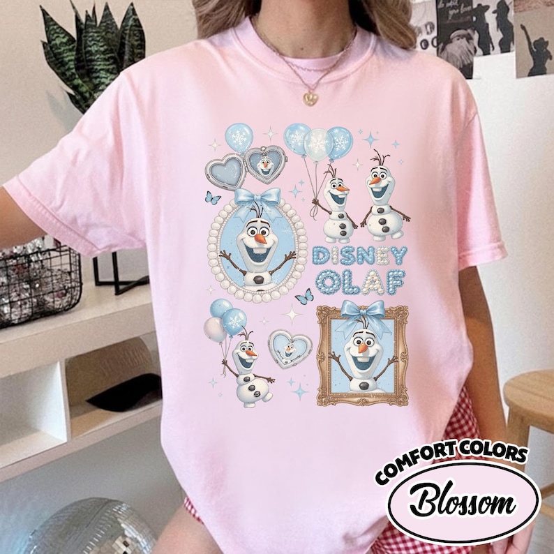 May include: Pink Comfort Colors t-shirt featuring a Disney Olaf graphic. The design includes Olaf characters, blue balloons, hearts, and the text "Disney Olaf". The shirt has a round neckline and short sleeves.