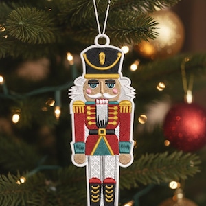 May include: Embroidered Christmas ornament shaped like a nutcracker soldier. The ornament features a red jacket, black hat, and white hair. It is hanging from a white string on a Christmas tree with gold and red ornaments.