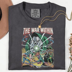 May include: Dark gray t-shirt featuring a comic book-style graphic with the text "THE WAR WITHIN." The design includes illustrations of superheroes and a villain. A gold necklace with a star pendant is draped over the shirt.