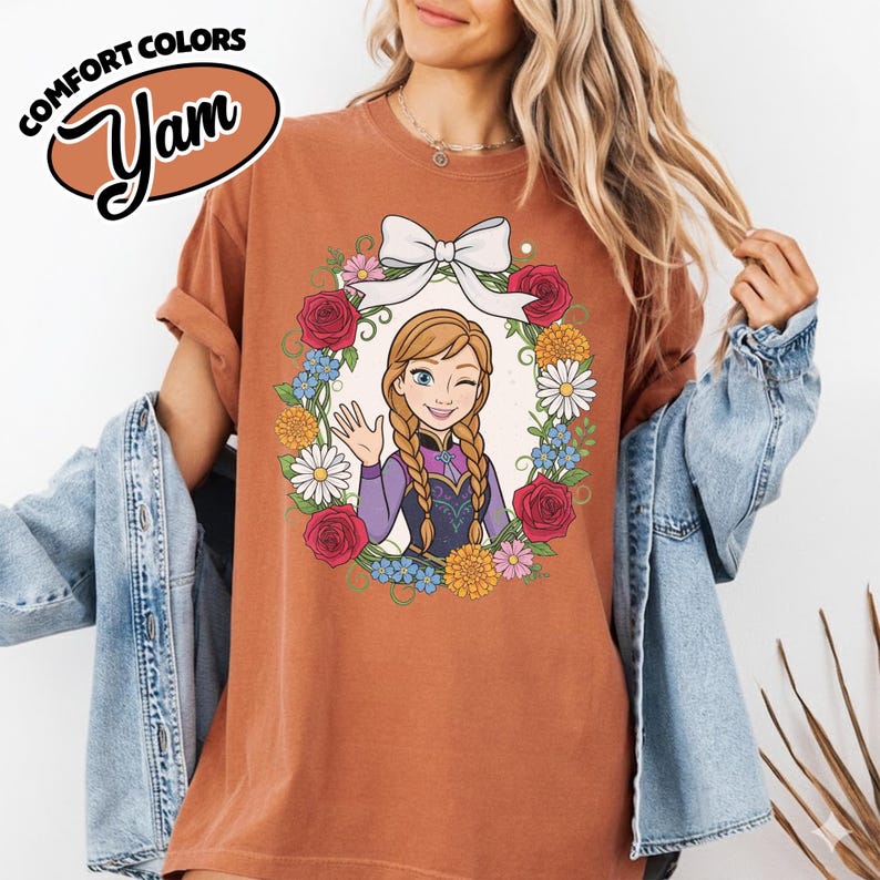 Peut inclure: T-shirt rouille avec un dessin anim&eacute; d'un personnage aux cheveux tress&eacute;s, faisant un clin d'&oelig;il et saluant, entour&eacute; d'une couronne florale et d'un n&oelig;ud blanc. Le t-shirt porte le texte "COMFORT COLORS" et "Yam" dans un ovale marron.