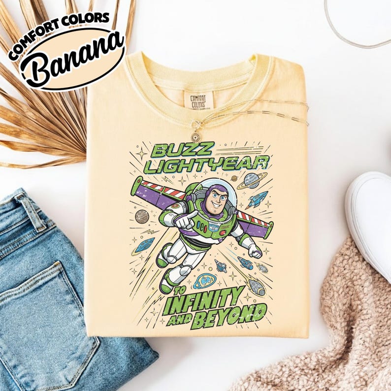 May include: A pale yellow t-shirt featuring a graphic of Buzz Lightyear in a space suit with wings extended. The text reads BUZZ LIGHTYEAR TO INFINITY AND BEYOND. The design includes small planets, stars, and rockets surrounding the character.