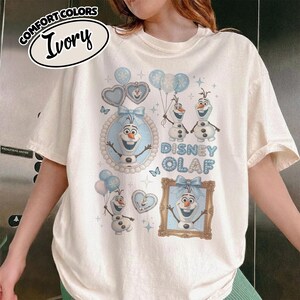 May include: Ivory-colored t-shirt featuring a Disney Olaf design. The design includes images of Olaf, hearts, balloons, and the words "Disney Olaf" in blue. The shirt has short sleeves and a crew neck.
