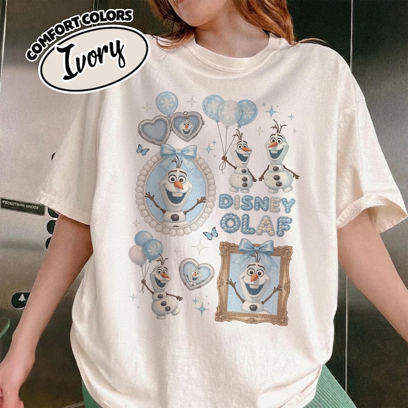 May include: Ivory-colored t-shirt featuring a Disney Olaf design. The design includes images of Olaf, hearts, balloons, and the words "Disney Olaf" in blue. The shirt has short sleeves and a crew neck.