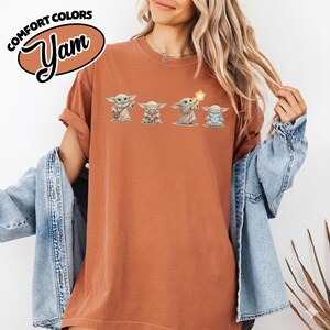 以下が含まれることがあります： 4つのグローグのイラストが描かれた、錆色のTシャツ。「COMFORT COLORS」と「Yam」の文字が茶色の楕円で描かれています。グローグのイラストは様々なポーズをしており、そのうちの1つは星を持っています。