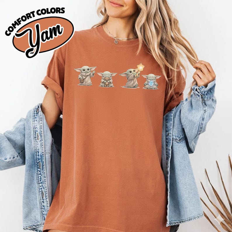 以下が含まれることがあります： 4つのグローグのイラストが描かれた、錆色のTシャツ。「COMFORT COLORS」と「Yam」の文字が茶色の楕円で描かれています。グローグのイラストは様々なポーズをしており、そのうちの1つは星を持っています。