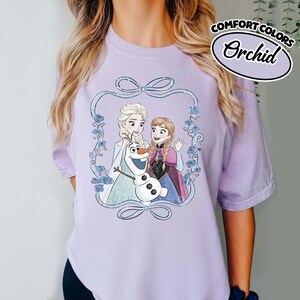 May include: A light purple t-shirt featuring a graphic of Elsa, Anna, and Olaf from the movie Frozen, framed by a blue ribbon and floral design. The shirt has the words "COMFORT COLORS" and "Orchid" printed on the upper right.