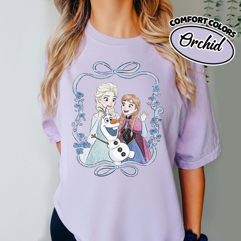 May include: A light purple t-shirt featuring a graphic of Elsa, Anna, and Olaf from the movie Frozen, framed by a blue ribbon and floral design. The shirt has the words "COMFORT COLORS" and "Orchid" printed on the upper right.