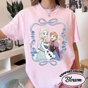May include: A light pink t-shirt featuring a graphic of Elsa, Anna, and Olaf from Frozen, framed by a blue ribbon and roses. The shirt has the words "Comfort Colors" and "Blossom" printed on it.