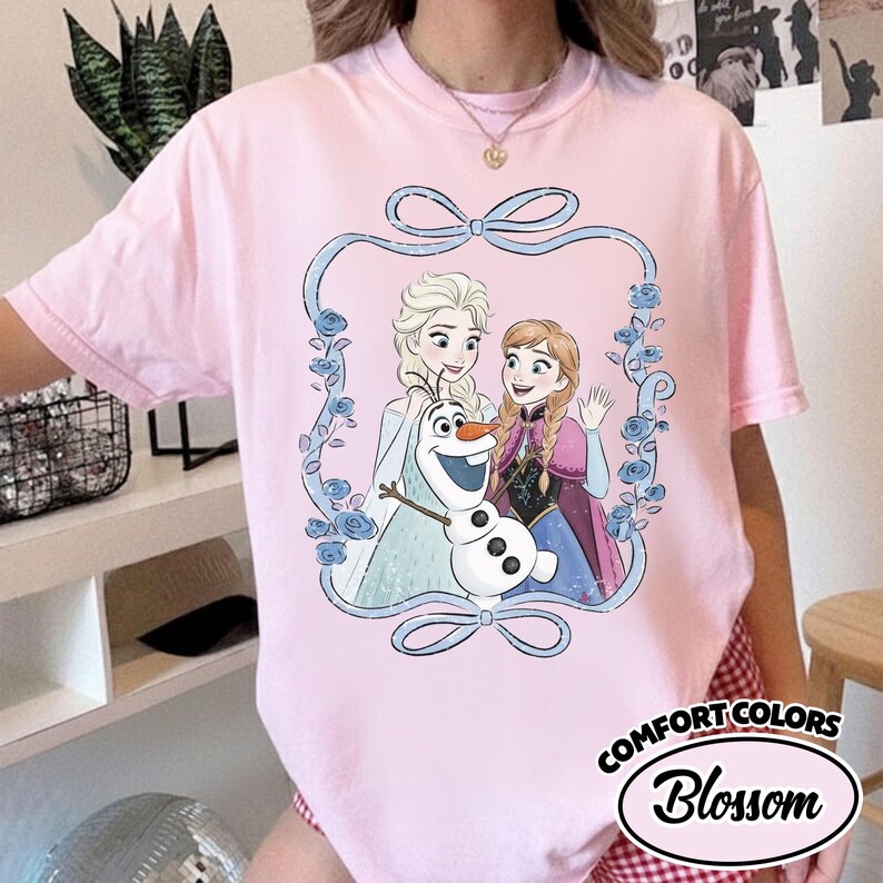 May include: A light pink t-shirt featuring a graphic of Elsa, Anna, and Olaf from Frozen, framed by a blue ribbon and roses. The shirt has the words "Comfort Colors" and "Blossom" printed on it.