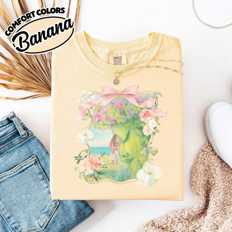 K&ouml;nnte beinhalten: Ein hellgelbes Comfort Colors Sweatshirt mit einer floralen und fig&uuml;rlichen Grafik. Die Grafik zeigt ein Frauengesicht mit einem Blumenkranz und einer rosa Schleife. Der Text "Banana" befindet sich in einem schwarzen Oval oben links.