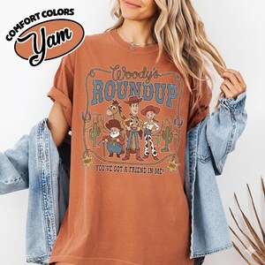 May include: A rust-colored t-shirt featuring a graphic of Woody's Roundup characters. The design includes Woody, Jessie, and other characters with the text "Woody's Roundup" and "You've Got a Friend in Me!". The shirt also has the Comfort Colors and Yam logos.