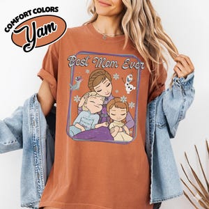 May include: A rust-colored t-shirt with the text "Best Mom Ever" and a cartoon illustration of a mother and two children. The shirt also features the Comfort Colors and Yam logos.