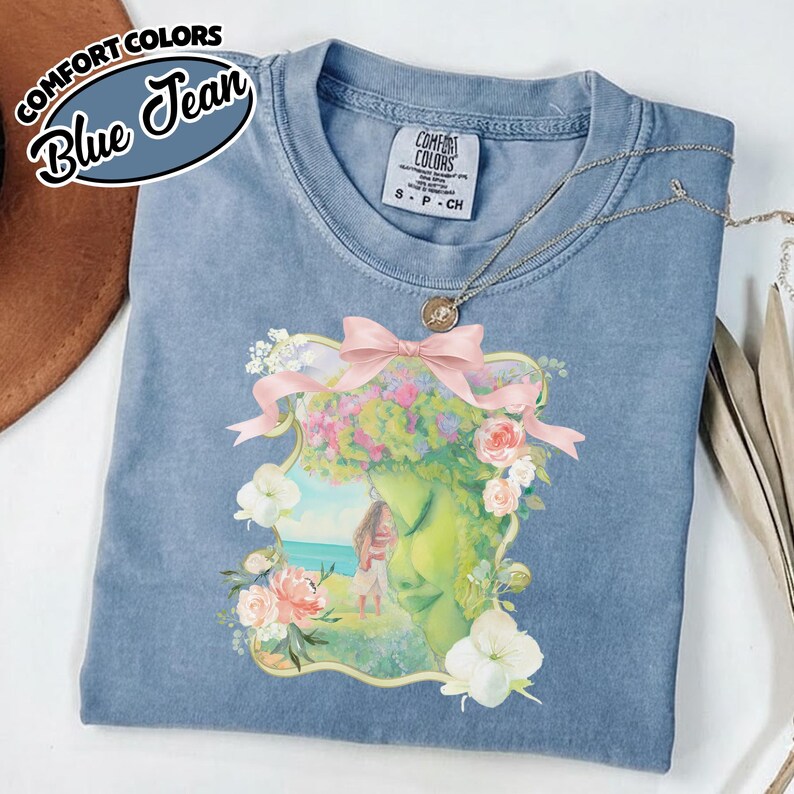 K&ouml;nnte beinhalten: Blaues Jeans-Comfort-Colors-T-Shirt mit floraler und fig&uuml;rlicher Grafik. Die Grafik zeigt ein Frauengesicht mit Blumen und einer rosa Schleife. Das Shirt hat einen Rundhalsausschnitt und kurze &Auml;rmel. Der Text "Comfort Colors" und "Blue Jean" befindet sich oben links.