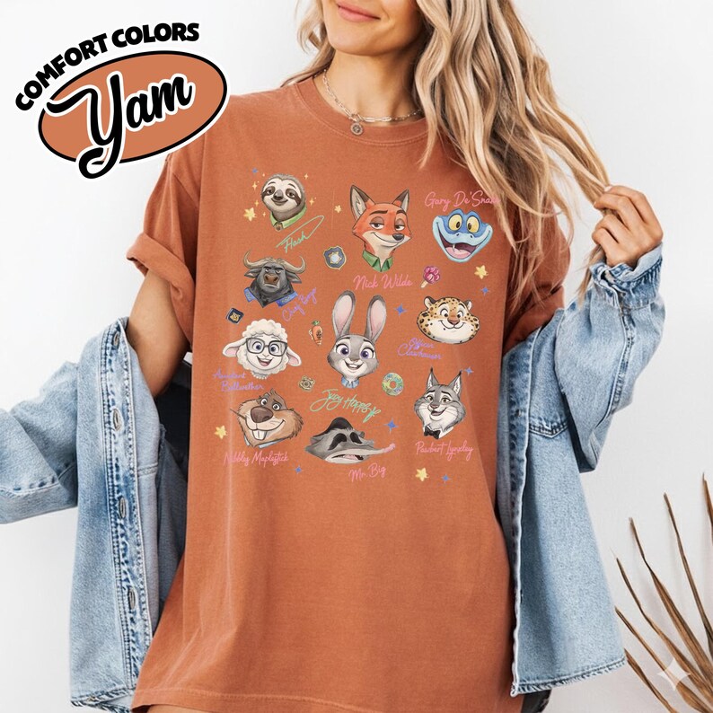K&ouml;nnte beinhalten: Ein rostfarbenes T-Shirt mit Illustrationen von Zeichentrickfiguren aus Zootopia. Auf dem Shirt stehen die W&ouml;rter "COMFORT COLORS" und "Yam" links oben. Die Illustrationen umfassen Judy Hopps, Nick Wilde und andere Charaktere.