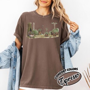 May include: Espresso-colored t-shirt featuring a cartoon scene with a character in a forest setting. The graphic includes multiple characters and the text "COMFORT COLORS Espresso". The shirt has rolled-up sleeves.