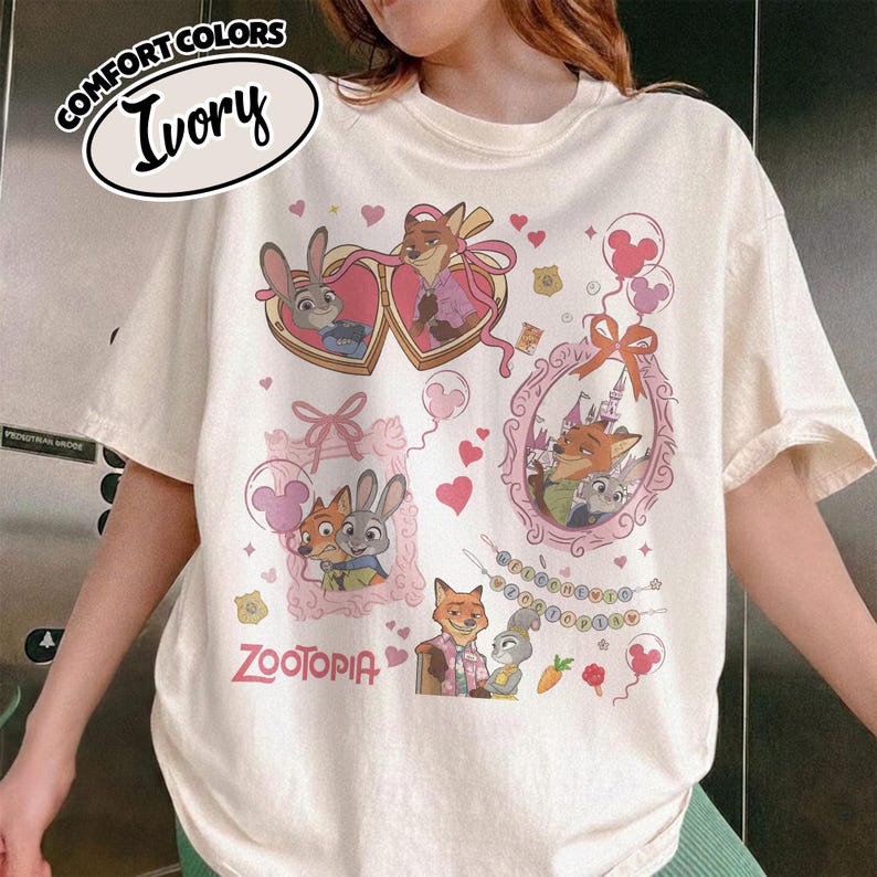 May include: An ivory t-shirt featuring a Zootopia graphic print with characters Judy Hopps and Nick Wilde. The design includes heart-shaped frames, pink bows, balloons, and the text Zootopia. A Comfort Colors Ivory label is visible in the top left corner.