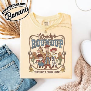 May include: A light yellow t-shirt with the text "Woody's Roundup" and a graphic of characters from the movie Toy Story. The shirt also has the text "You've Got a Friend in Me!" and the words "Comfort Colors" and "Banana".