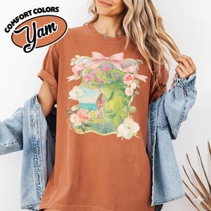 K&ouml;nnte beinhalten: Ein rostfarbenes T-Shirt mit einem floralen Grafikdesign. Die Grafik zeigt ein Frauengesicht mit Blumenkrone, einer rosa Schleife und einer Landschaftsszene. Die Worte "COMFORT COLORS" und "Yam" sind oben links aufgedruckt.