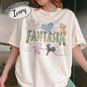 May include: Ivory-coloured t-shirt with the word "Fantasia" in green letters, and cartoon pegasus characters and clouds. The shirt has a relaxed fit and the words "Comfort Colors" and "Ivory" are printed on the top left.