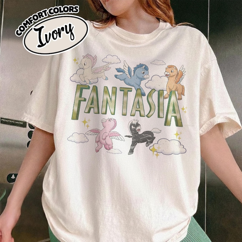 May include: Ivory-coloured t-shirt with the word "Fantasia" in green letters, and cartoon pegasus characters and clouds. The shirt has a relaxed fit and the words "Comfort Colors" and "Ivory" are printed on the top left.