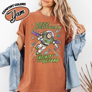 May include: A rust-colored short-sleeve t-shirt featuring a graphic of Buzz Lightyear flying through space. The design includes the text "BUZZ LIGHTYEAR" and "TO INFINITY AND BEYOND" in green, surrounded by planets, stars, and small spacecraft illustrations.