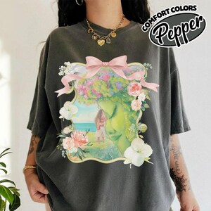 K&ouml;nnte beinhalten: Dunkelgraues T-Shirt mit floralem Design, das ein Frauengesicht, Blumen und eine rosa Schleife zeigt. Das Shirt hat den Text "COMFORT COLORS Pepper" in einem wei&szlig;en und schwarzen Oval.