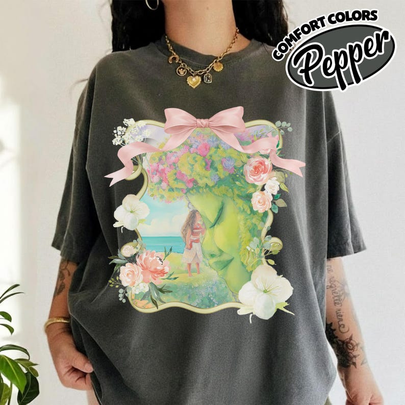 K&ouml;nnte beinhalten: Dunkelgraues T-Shirt mit floralem Design, das ein Frauengesicht, Blumen und eine rosa Schleife zeigt. Das Shirt hat den Text "COMFORT COLORS Pepper" in einem wei&szlig;en und schwarzen Oval.