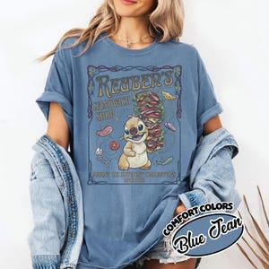 K&ouml;nnte beinhalten: Blaues Jeans Comfort Colors T-Shirt mit einer Cartoon-Illustration einer Figur, die ein gro&szlig;es Sandwich h&auml;lt. Das Shirt hat den Text "Reuber's Sandwich Shop" und "Making 125 Different Combinations Since 2002".