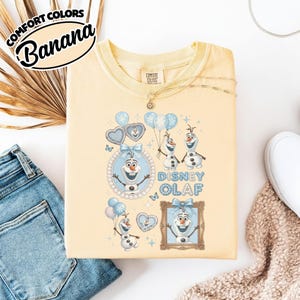 May include: A light yellow sweatshirt featuring the text "Comfort Colors Banana" and a Disney Olaf graphic. The graphic includes Olaf the snowman, balloons, and the words "Disney Olaf" in blue. The sweatshirt is paired with blue jeans and a necklace.