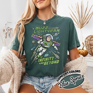 May include: A dark teal short-sleeve t-shirt featuring a graphic of Buzz Lightyear flying through space. The text reads BUZZ LIGHTYEAR TO INFINITY AND BEYOND. The design includes planets, stars, and a comet, rendered in shades of green, purple, and white.