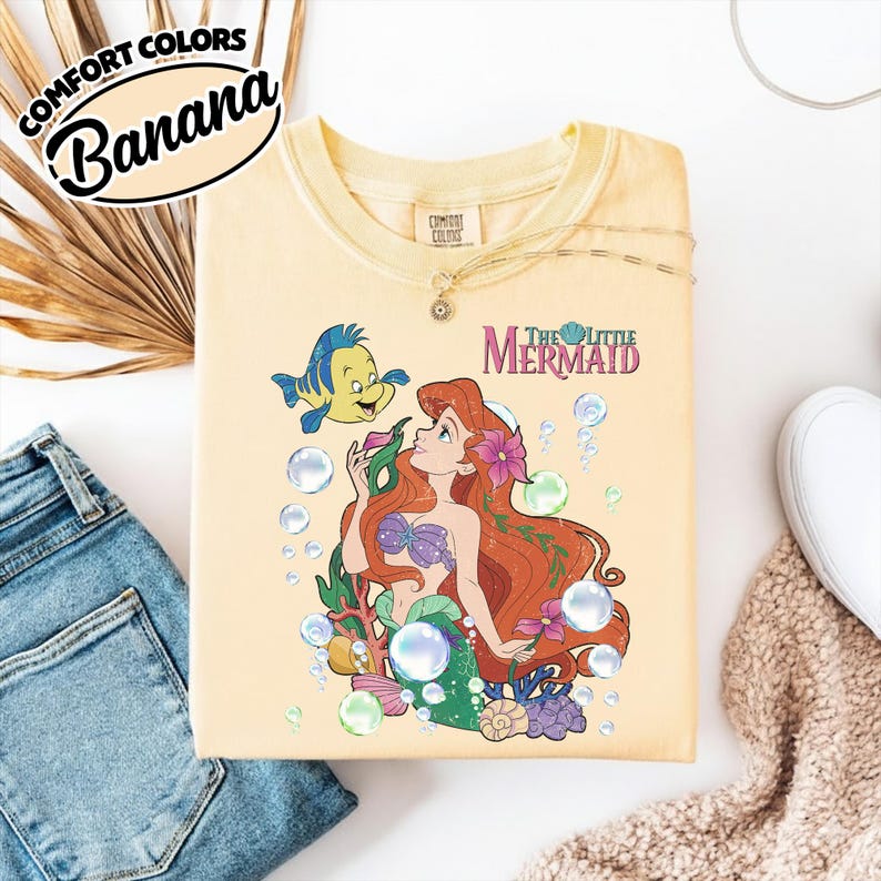 K&ouml;nnte beinhalten: Ein blassgelbes T-Shirt mit einem Grafikdruck von Arielle und Fabius aus Arielle, die Meerjungfrau. Das Design enth&auml;lt bunte Unterwasserelemente wie Blasen, Seetang und Muscheln, wobei der Text The Little Mermaid in Lila aufgedruckt ist.