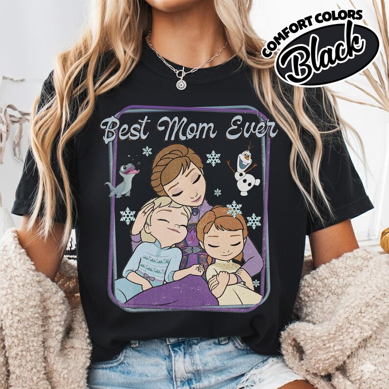 May include: Black t-shirt with a graphic featuring the phrase "Best Mom Ever" and an illustration of a mother and two children, with snowflakes and cartoon characters. The shirt has a Comfort Colors label.
