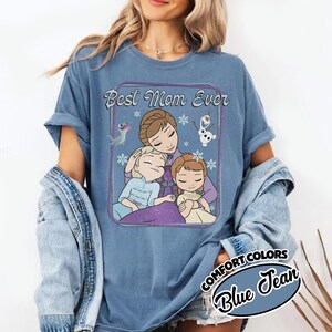 May include: Blue Comfort Colors t-shirt with a graphic featuring the text "Best Mom Ever" and an illustration of a mother and two children, with snowflakes and cartoon characters. The shirt is a light blue color.