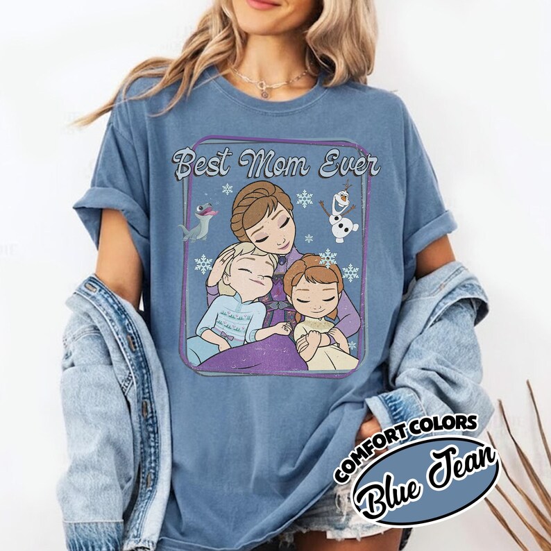 May include: Blue Comfort Colors t-shirt with a graphic featuring the text "Best Mom Ever" and an illustration of a mother and two children, with snowflakes and cartoon characters. The shirt is a light blue color.