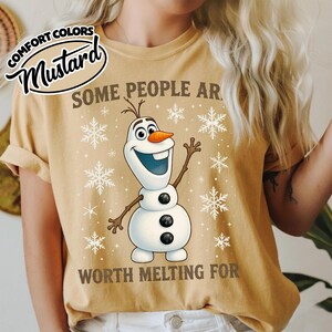 May include: Mustard-colored t-shirt featuring Olaf the snowman from Frozen, surrounded by snowflakes. The text reads "SOME PEOPLE ARE WORTH MELTING FOR". The shirt has rolled-up sleeves and a Comfort Colors label.