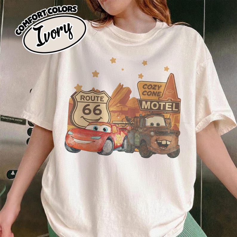 May include: Ivory-colored t-shirt featuring a graphic of characters from the movie Cars, including Lightning McQueen and Mater, with Route 66 and Cozy Cone Motel signs. The shirt has the words "Comfort Colors" and "Ivory" printed on it.