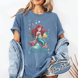 May include: Blue jean Comfort Colors t-shirt featuring a graphic of Ariel from The Little Mermaid. The graphic includes Ariel, Flounder, Sebastian, and other sea elements. The text "THE LITTLE MERMAID" is below the graphic.