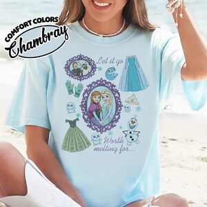 Op de afbeelding: Lichtblauw T-shirt met een grafische afbeelding met personages en elementen uit een populaire animatiefilm. Het ontwerp bevat de tekst "Let it go" en "Worth melting for..." samen met illustraties van een jurk, handschoenen en sneeuwvlokken.