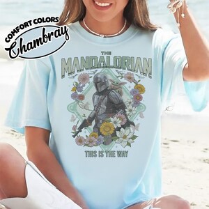 May include: A light blue t-shirt featuring a graphic of The Mandalorian and Grogu surrounded by colorful flowers and butterflies. The text reads THE MANDALORIAN at the top and THIS IS THE WAY at the bottom. A Comfort Colors Chambray logo is visible.