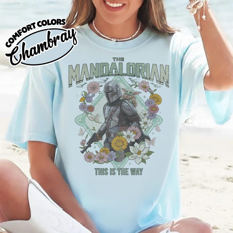 May include: A light blue t-shirt featuring a graphic of The Mandalorian and Grogu surrounded by colorful flowers and butterflies. The text reads THE MANDALORIAN at the top and THIS IS THE WAY at the bottom. A Comfort Colors Chambray logo is visible.