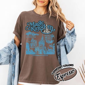 May include: Brown t-shirt with a blue graphic that says "Magic Kingdom" and features Cinderella's castle, Autopia car, Peter Pan's Flight vehicle, and other park attractions. The shirt is labeled "Comfort Colors Espresso".