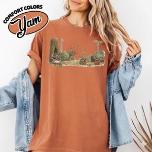 May include: Rust-colored t-shirt with a graphic of a forest scene featuring a character from a popular science fiction series. The graphic includes the text "Comfort Colors" and "Yam" in a brown oval. The shirt has rolled-up sleeves.