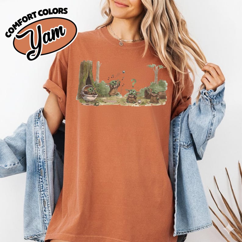 May include: Rust-colored t-shirt with a graphic of a forest scene featuring a character from a popular science fiction series. The graphic includes the text "Comfort Colors" and "Yam" in a brown oval. The shirt has rolled-up sleeves.