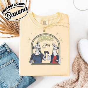 May include: A pale yellow t-shirt featuring the text "ZEBROS" and a graphic of two zebras. One zebra is in overalls, and the other in a jacket. The shirt also has the text "Comfort Colors Banana" in a circular design.