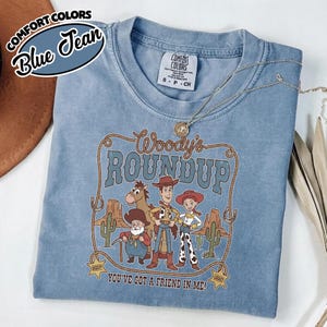 May include: A blue jean-colored t-shirt featuring the text "Woody's Roundup" with cartoon characters. The shirt has a crew neck and short sleeves. The text "You've Got a Friend in Me!" is printed at the bottom.