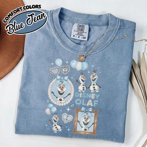 May include: Blue jean Comfort Colors t-shirt featuring Disney's Olaf. The shirt has a crew neck and short sleeves. The graphic includes Olaf in various poses, hearts, and balloons. The text "Disney Olaf" is also visible. A silver necklace is draped across the shirt.